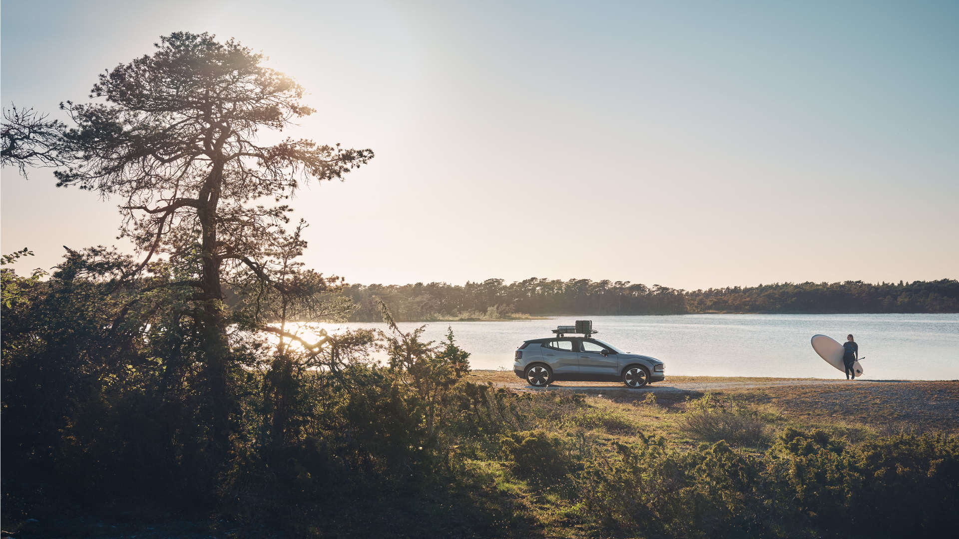 Overview-location-gallery-16x9 SUV med takboks parkert ved innsjø – person med paddleboard forbereder seg på aktivitet i solnedgang