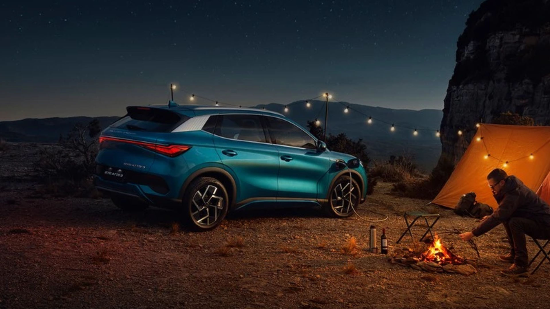 BYD Atto 3 i blå farge parkert ved et telt i fjellandskap om natten, med bilen koblet til lading og et bål ved siden av.