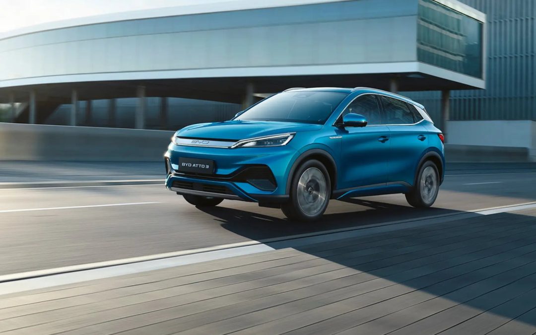 Blå BYD Atto 3 i bevegelse på en moderne vei, med et stilrent bygg i bakgrunnen.