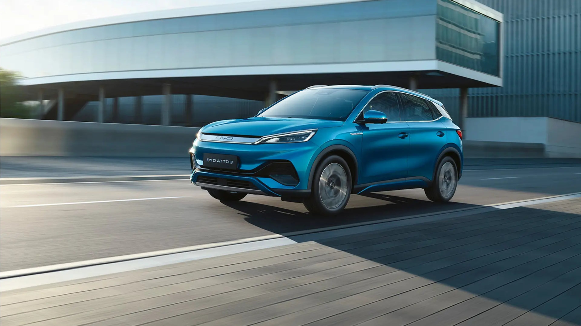 Blå BYD Atto 3 i bevegelse på en moderne vei, med et stilrent bygg i bakgrunnen.